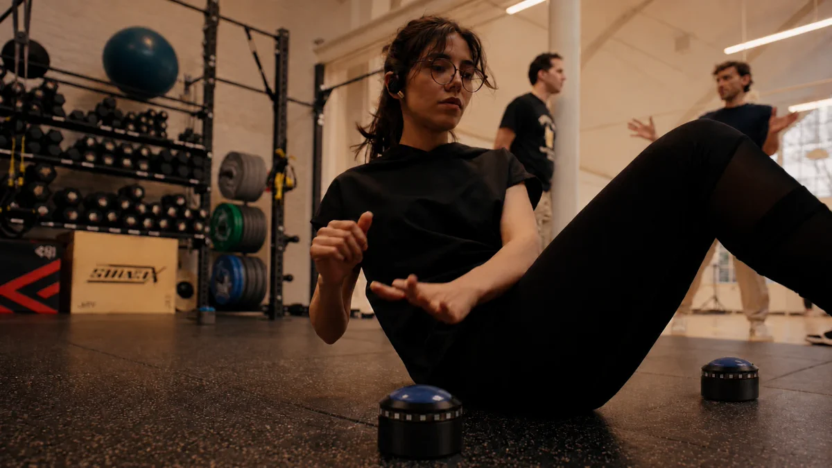 A participant seated on the gym floor between two Flux Pods reaches towards one of them, wearing glasses and bone-conduction headphones, while two people talk behind her.